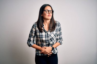 Mavi gözlü genç esmer kadın günlük gömlek ve gözlük takıyor beyaz arka plan üzerine el üstünde midesi çünkü hazımsızlık, acı verici bir hastalık kendini iyi hissetmiyor. Ache kavramı.