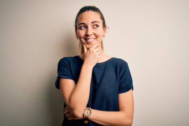 Mavi gözlü, genç, güzel, sarışın bir kadın beyaz arka planda sıradan tişörtler giyiyor ve elleri çenesinde soru sormayı düşünüyor. Düşünceli bir yüz ifadesi var. Düşünceli bir yüzle gülümsüyor. Şüphe kavramı.