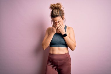 Genç ve güzel sarışın sporcu kadın spor yapıyor. Pembe arka planda spor kıyafetleri giyiyor. Yorgunluk ve baş ağrısı için gözlerini ovuşturuyor. Görüş sorunu