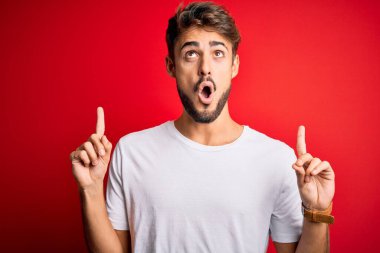 Kırmızı arka plan üzerinde duran gündelik tişört giyen sakallı yakışıklı genç adam şaşırmış ve şaşırmış bir şekilde yukarı bakıp parmaklarını göstermiş ve kollarını kaldırmıştı..
