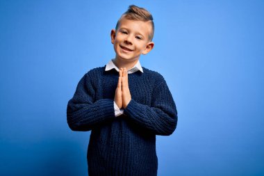 Mavi gözlü küçük beyaz çocuk mavi arka plan üzerine kışlık süveter giyip ellerinin birarada dua ederek af diliyor kendinden emin bir şekilde gülümsüyor..
