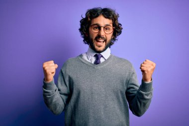 Sakallı, kravat takan ve mor arka planda duran yakışıklı iş adamı kollarını kaldırarak ve açık gözlerle başarıyı kutluyor ve şaşırıyor. Kazanan konsept.