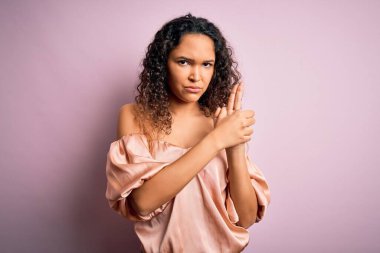 Kıvırcık saçlı, günlük tişörtlü pembe arka planda duran, el hareketleriyle sembolik silah tutan, cinayet silahlarıyla oynayan, kızgın yüzlü genç ve güzel bir kadın.