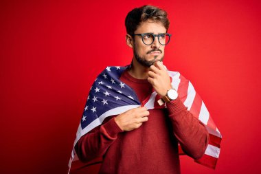 Gözlüklü genç adam ve Amerika Birleşik Devletleri bayrağı izole edilmiş kırmızı arka plan üzerinde ciddi bir yüz soru düşünüyor, çok karışık bir fikir.