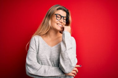 İzole edilmiş kırmızı arka plan üzerinde süveter ve gözlük takan genç ve güzel sarışın kadın kameraya kollarını kavuşturup gülümseyerek ve çenesini kaldırarak bakıyor. Olumlu düşünüyorum..