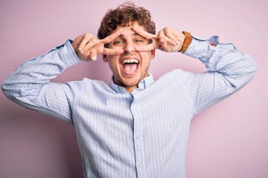 Sarışın, kıvırcık saçlı, beyaz arka planda çizgili gömlek giyen, yüzünde parmaklarıyla barış sembolü çizen, neşeyle gülümseyen, zaferi gösteren genç bir adam.