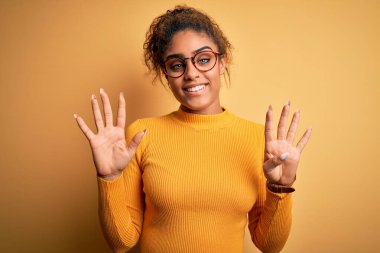 Sarı arka planda süveter ve gözlük takan güzel Afro-Amerikan kızı dokuz numaralı parmaklarıyla işaret ederken kendinden emin ve mutlu gülümsüyor..
