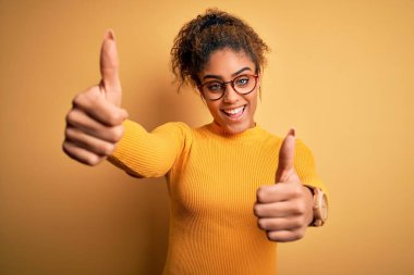Sarı arka planda süveter ve gözlük takan güzel afro-amerikalı kız el hareketlerini onaylıyor, gülümsüyor ve başarı için mutlu. Kazanan jest.