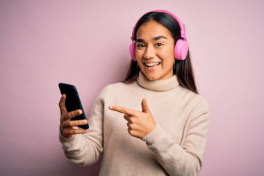 Genç ve güzel Asyalı kadın akıllı telefon ve kulaklık kullanarak müzik dinliyor. El ve parmak göstererek çok mutlu oluyorlar.