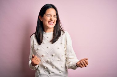 Mavi gözlü genç esmer kadın izole edilmiş pembe arka plan üzerine günlük süveter giyiyor. Kolları havada ve gözleri kapalı zaferi kutlarken gülümsüyor. Kazanan konsept.