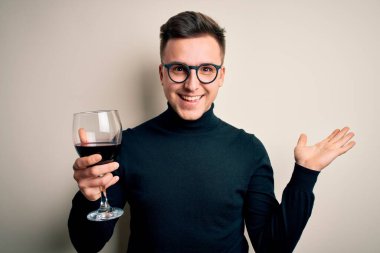 Genç beyaz adam izole edilmiş arka planda alkollü bir bardak kırmızı şarap içiyor mutlu ve heyecanlı, kazanan ifade zaferi büyük bir gülümsemeyle kutluyor ve ellerini kaldırıyor.