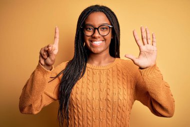 Genç Afro-Amerikan akıllı kadın gözlük takıyor ve sarı arka planda rahat bir süveter giyiyor. Altıncı parmaklarıyla işaret ediyor ve gülümsüyor..