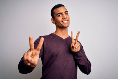 Beyaz arka planda rahat süveter giyen yakışıklı Afro-Amerikalı genç adam kameraya bakıp zafer işareti yapıyor. İki numara..