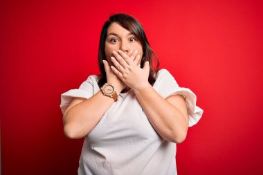 Güzel esmer ve uzun boylu bir kadın izole edilmiş kırmızı arka plan üzerine gündelik tişört giyerek ağzını yanlışlıkla ellerle kapatmış. Gizli konsept.