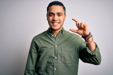 İzole edilmiş beyaz arka plan üzerinde dikilen Brezilyalı genç adam gülümsüyor ve el hareketleriyle küçük beden işareti yaparak kameraya bakıyor. Ölçü kavramı.