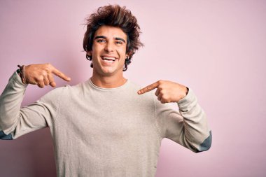 İzole edilmiş pembe arka planda duran sıradan tişört giyen genç yakışıklı adam yüzünde bir gülümsemeyle gururlu ve mutlu bir şekilde kendini işaret ediyor..