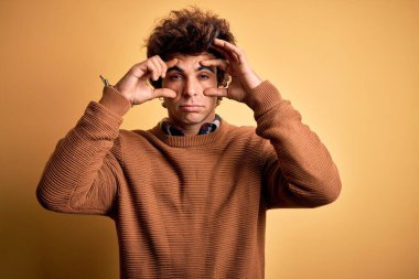 Sarı arka plan üzerinde gündelik gömlek ve süveter giyen genç yakışıklı adam gözlerini parmaklarıyla açmaya çalışıyor, uykulu ve yorgun bir şekilde sabah yorgunluğu çekiyor.