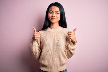 Young beautiful chinese woman wearing casual sweater over isolated pink background success sign doing positive gesture with hand, thumbs up smiling and happy. Cheerful expression and winner gesture.