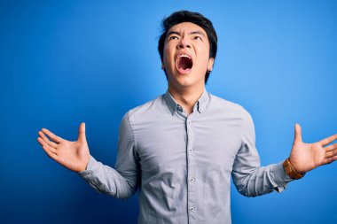 Genç, yakışıklı Çinli bir adam, günlük gömleğini giymiş, mavi arka planda dikiliyor, deli deli deli bağırıyor, agresif bir ifadeyle bağırıyor ve kollarını kaldırıyor. Hayal kırıklığı konsepti.