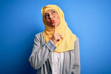Orta yaşlı esmer iş kadını, geleneksel Müslüman tesettüre bürünmüş mavi arka planda şüpheye odaklanmış, parmakları çenesinde ve yukarı bakıyor, merak içinde.