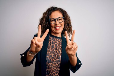 Orta yaşlı esmer iş kadını, gözlüklü, izole edilmiş beyaz arka planda gülümseyen, parmaklarını zafer işareti yaparken gösteren kameraya bakan. İki numara..