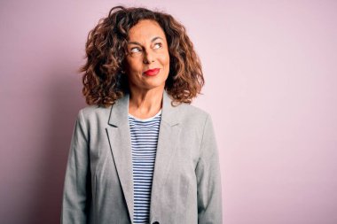 Orta yaşlı, güzel bir iş kadını, zarif bir ceket giyiyor, pembe arka planda. Gülümsüyor ve uzaklara bakıyor..