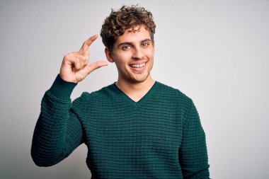 Sarışın, kıvırcık saçlı, yeşil süveter giyen, beyaz arka planda gülümseyen, kendine güvenen, el hareketleriyle küçük boy işareti yapan, parmaklarıyla işaret eden ve kameraya bakan genç bir adam. Ölçü kavramı.