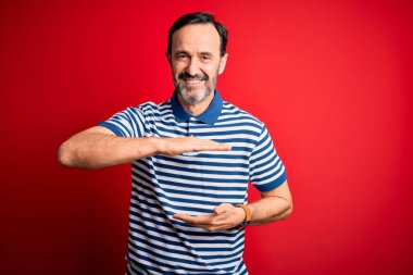 Orta yaşlı, beyaz, sıradan çizgili polo giyen, izole edilmiş kırmızı arka plan üzerinde duran, büyük ve büyük boyut işareti gösteren, ölçü sembolü olan bir adam. Kameraya bakıp gülümsüyor. Ölçüm konsepti.