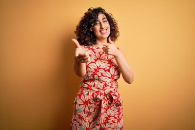 Genç ve güzel kıvırcık Arap kadın tatilde. Çiçekli bir elbise giyiyor ve güneş gözlüğü takıyor. Mutlu ve komik yüzüyle kamerayı işaret ediyor. İyi enerji ve titreşimler.