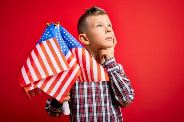 Küçük Amerikalı vatansever beyaz çocuk kırmızı arka planda ABD bayrağı taşıyor. Ciddi bir yüz. Soru üzerine kafa karıştırıcı bir düşünce.