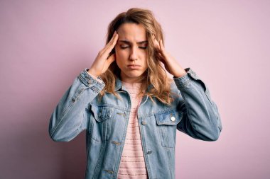 Genç, güzel, sarışın bir kadın. Günlük kot ceket giyiyor. Pembe arka planda duruyor. Migren ağrısı..
