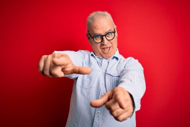 Orta yaşlı yakışıklı, beyaz gömlekli ve gözlüklü, kırmızı arka planda seni ve kamerayı işaret eden neşeli ve pozitif gülümseyen bir adam.