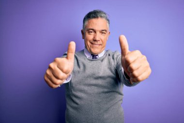 Orta yaşlı, yakışıklı, gri saçlı, mor arka planda zarif bir süveter giyen, elle olumlu bir jest yapmayı onaylayan, gülümseyen ve başarı için mutlu bir adam. Kazanan jest.