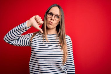 Kırmızı arka planda duran gözlüklü, mavi gözlü, genç, güzel, sarışın bir kadın. Mutsuz ve sinirli görünüyor. Reddedildiğini gösteriyor. Kötü ifade.