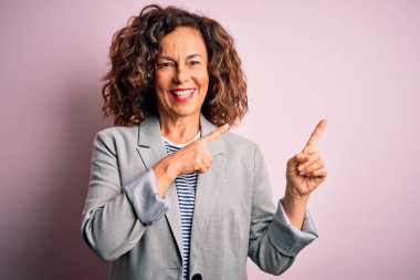 Orta yaşlı, güzel iş kadını zarif bir ceket giyiyor pembe arka planda gülümsüyor ve kameraya bakıyor iki eliyle ve parmaklarıyla yan tarafı işaret ediyor..