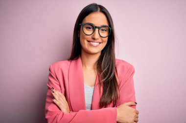 Güzel, esmer bir iş kadını pembe arka planda gözlük takıp kollarını kavuşturup kameraya bakarken gülümsüyor. Pozitif kişi.
