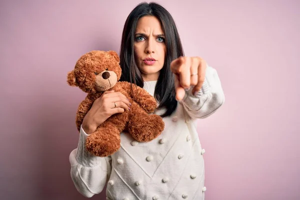 Young brunette woman with blue eyes hugging teddy bear stuffed animal over pink background pointing with finger to the camera and to you, hand sign, positive and confident gesture from the front