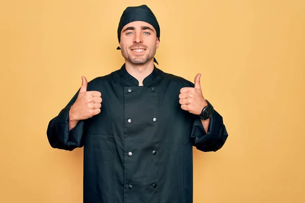 年轻英俊的炊事员 蓝眼睛 身穿制服 头戴黄底帽 手握大拇指 微笑着 快乐地做着积极的手势 快乐的表情和胜利的姿态 — 图库照片
