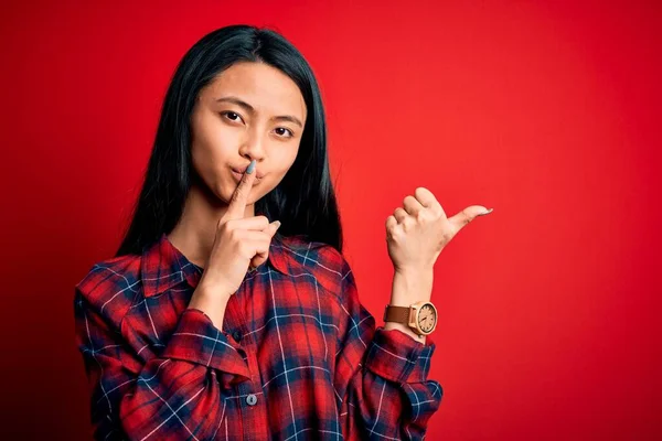 年轻美丽的中国女人穿着宽松的衬衫 披着孤立的红色背景 要求保持安静 手指放在嘴唇上 手拉手放在侧面 沉默和秘密概念 — 图库照片