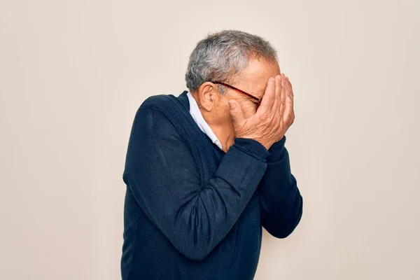 Senior handsome grey-haired man wearing sweater and glasses over isolated white background with sad expression covering face with hands while crying. Depression concept.