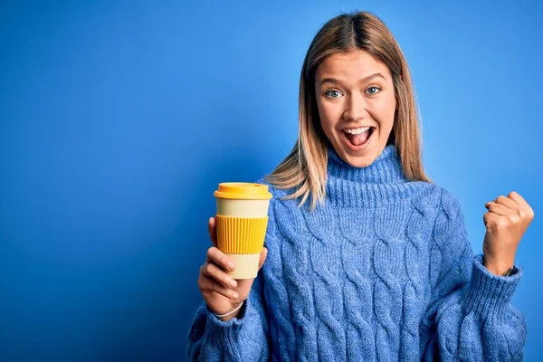 Young beautiful woman holding take away glass of coffee over isolated blue background screaming proud and celebrating victory and success very excited, cheering emotion