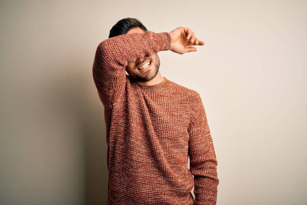 Young handsome man wearing casual sweater standing over isolated white background covering eyes with arm smiling cheerful and funny. Blind concept.