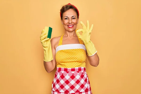 Middle age senior housewife pin up woman wearing 50s style retro dress using cleaner scrub doing ok sign with fingers, excellent symbol