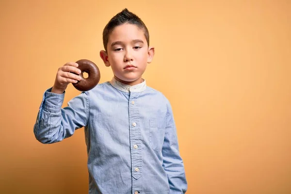 若いです男の子食べる不健康なチョコレートドーナツ上の隔離された黄色の背景とともに自信を持って式上のスマート顔考え深刻な — ストック写真
