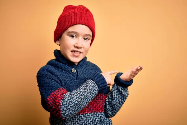 若いです男の子身に着けているウールキャップと冬のセーター上に黄色隔離された背景驚きと笑顔にカメラを提示しながら手と指差し指 — ストック写真