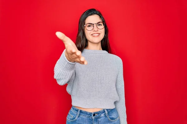 赤の背景にカジュアルなセーターを着た若い美しいブルネットの女性が挨拶と歓迎として握手を提供して笑顔 成功事業 — ストック写真