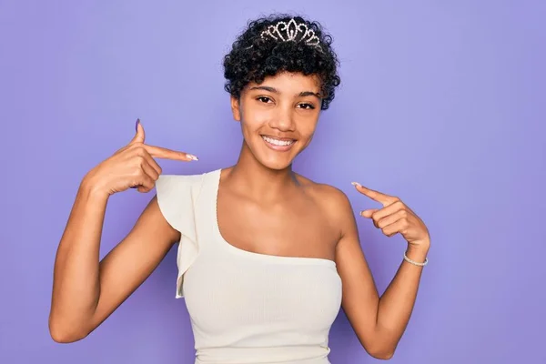 若い美しいアフリカ系アメリカ人のアフロ女性は 顔に笑顔で自信を持って見える紫色の背景にティアラ冠を身に着けています 誇りと幸せ指で自分自身を指して — ストック写真