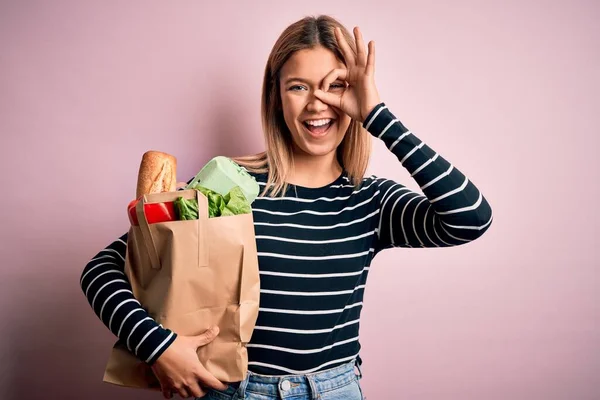 Young beautiful woman holding paper bag with purchase over isolated pink background with happy face smiling doing ok sign with hand on eye looking through fingers