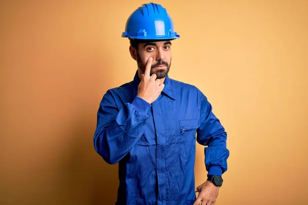 Mechanic man with beard wearing blue uniform and safety helmet over yellow background Pointing to the eye watching you gesture, suspicious expression