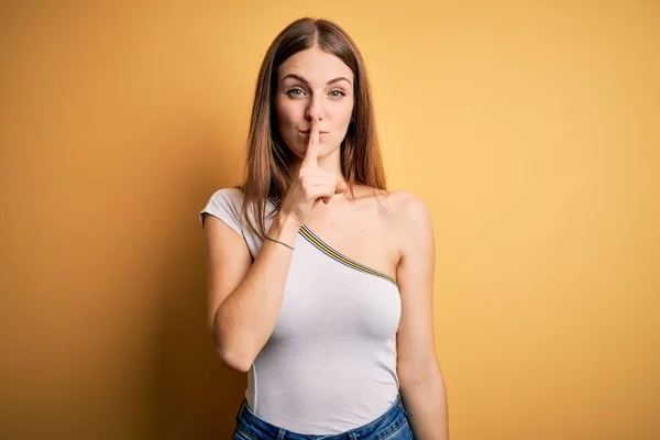 若い美しい赤毛の女性は 孤立した黄色の背景の上にカジュアルなTシャツを着て唇に指で静かにするように求めている 沈黙と秘密の概念 — ストック写真